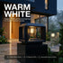 A modern black outdoor bollard light with a warm white illuminated glass bulb, mounted on a dark stone base in front of a contemporary house at dusk. Large text reads 'WARM WHITE' with additional product callouts on the right and bottom.