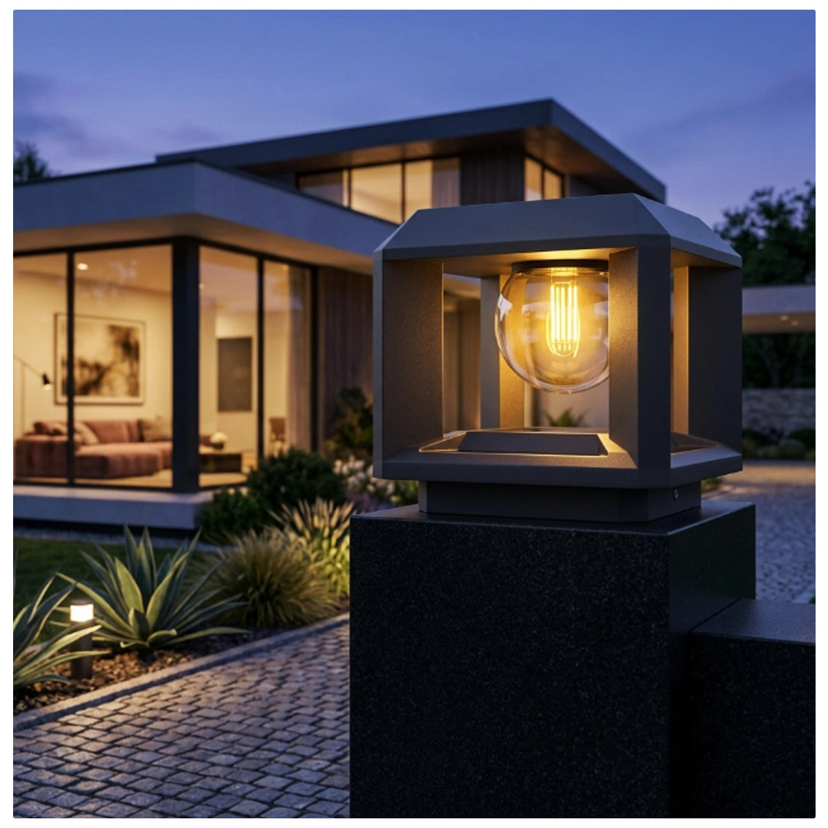 A modern outdoor bollard light with a dark gray metal frame and a glowing warm Edison bulb inside a clear glass shade, mounted on a dark post in front of a contemporary house at dusk.