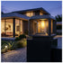 A modern outdoor bollard light with a dark gray metal frame and a glowing warm Edison bulb inside a clear glass shade, mounted on a dark post in front of a contemporary house at dusk.