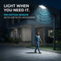 A modern outdoor LED street light with a black pole is shown at night beside a house, illuminating a walkway. A man is walking under the light, and blue graphic wave lines indicate motion detection.